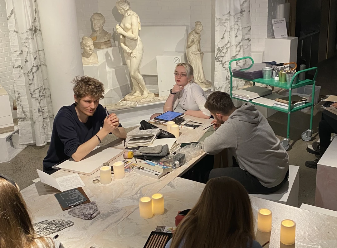 In a room with marble statues, people are gathered around tables with sketchbooks and paper. Bastian is explaining the drawing activity of the day.